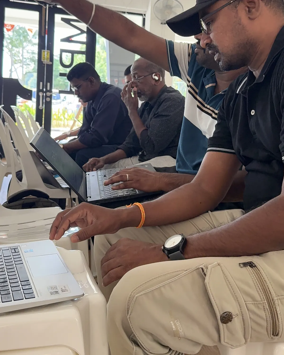 A learner in a cap points across the room while typing on a laptop, with others working behind